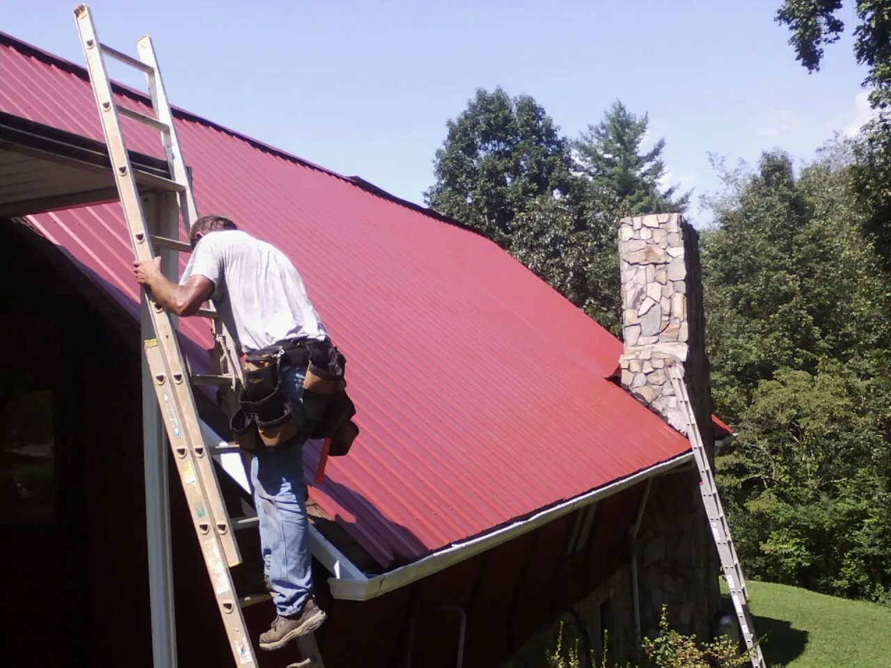Roofer on a ladder beside a red metal roof and stone chimney — Soffit & Fascia Repair by Roofing Brothers in Rush Valley